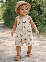 Smiling toddler in a bird-patterned romper and straw hat, standing on a gravel path surrounded by greenery.