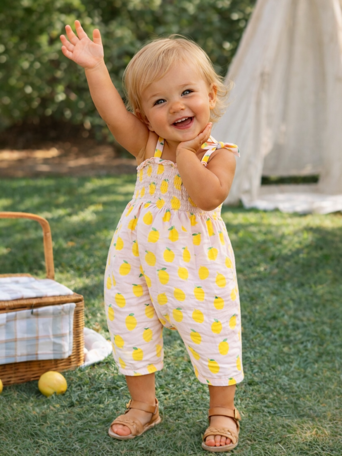 Toddler in lemon-patterned jumpsuit waves happily, standing on green grass with a picnic basket nearby.