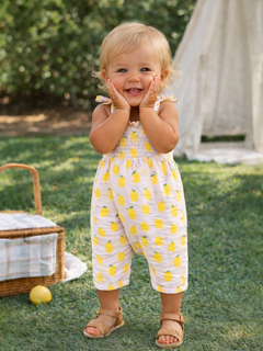 Smiling toddler in lemon-patterned outfit, hands on cheeks, standing in grassy area with a picnic basket nearby.