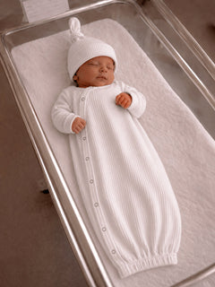 Newborn baby sleeping in a white ribbed gown and hat in a hospital bassinet.