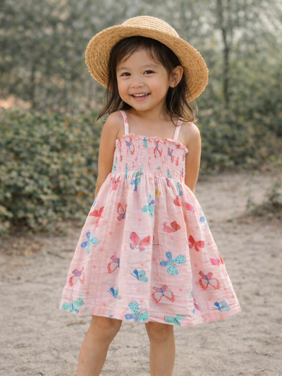 Smiling girl in a butterfly-patterned dress and straw hat, standing outdoors on a sunny day.