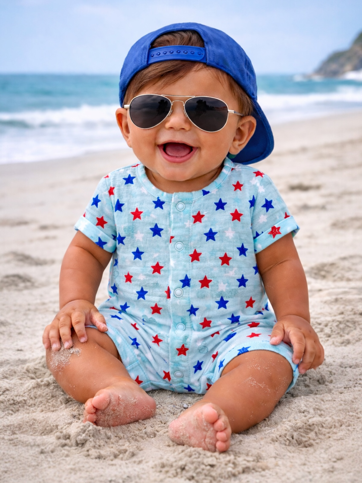 Smiling baby in star-patterned romper, wearing sunglasses and a blue cap, playing on a sandy beach.