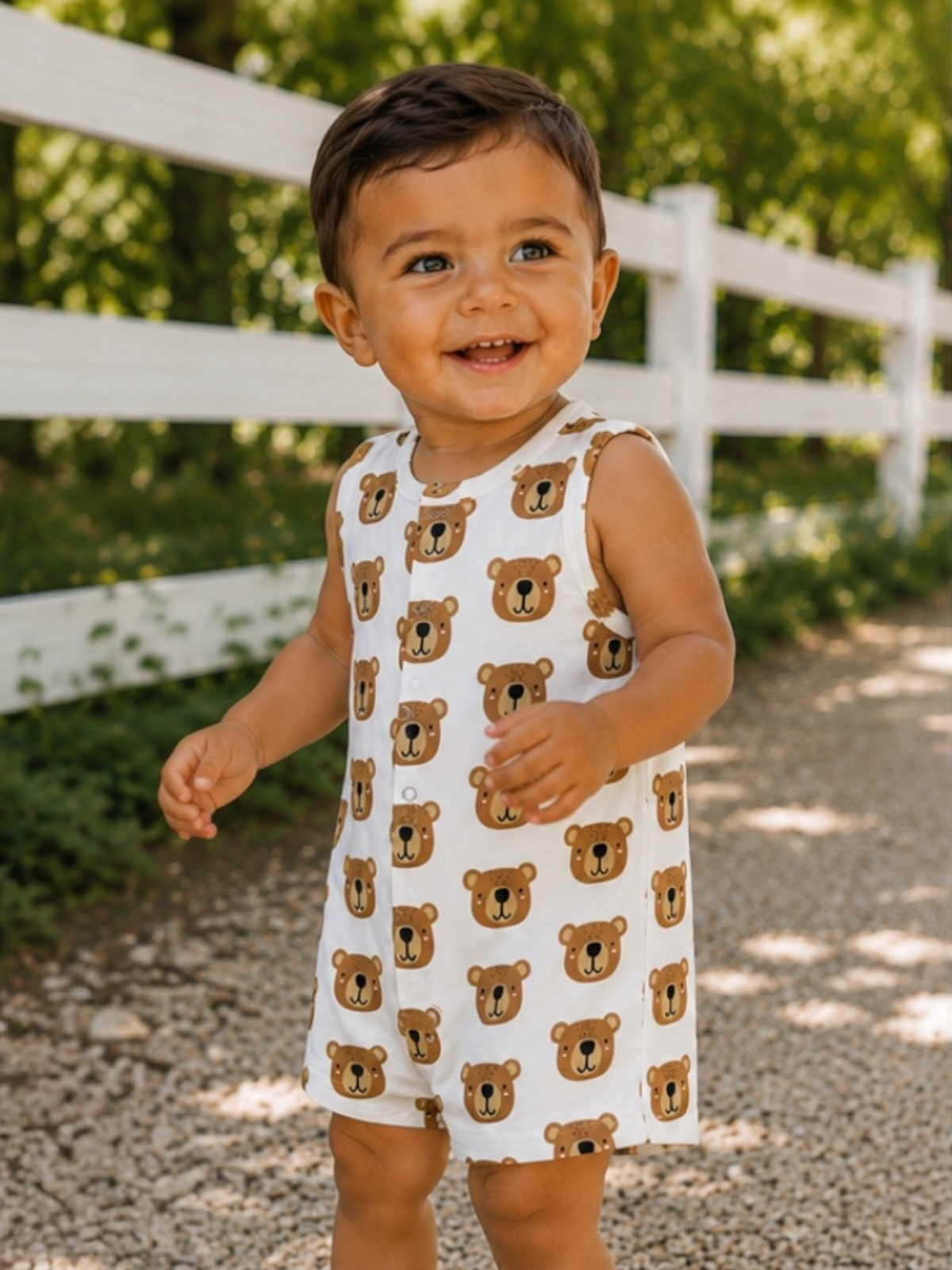 Smiling toddler in a bear-print outfit, playing outdoors near a white fence and gravel path, surrounded by greenery.