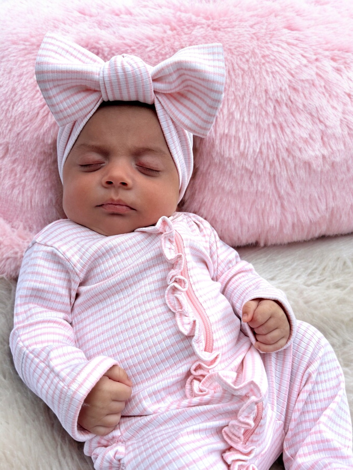 Sleeping baby in a pink outfit with a large bow, resting on a fluffy pink surface.