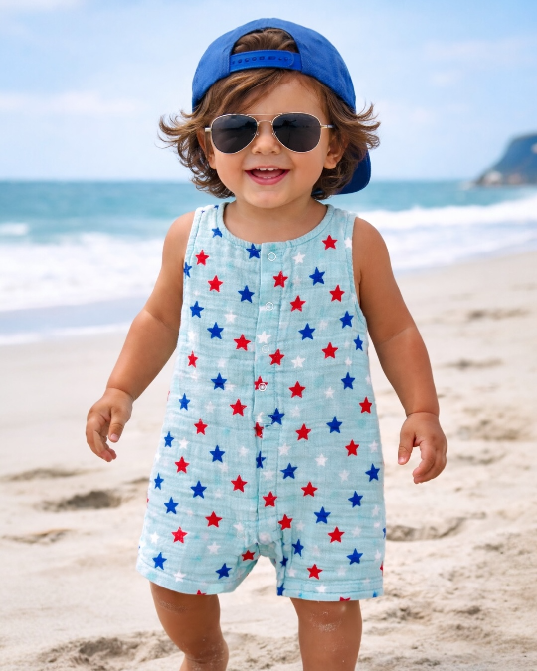Smiling child in sunglasses and a cap, wearing a star-patterned romper on a sunny beach.