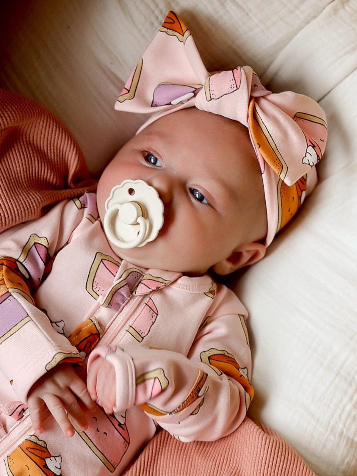 Baby in a pink outfit with dessert patterns and a large bow, lying on a soft surface, sucking a pacifier.