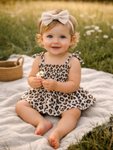 Smiling baby girl in a leopard-print dress holds a daisy, sitting on a blanket in a grassy field.