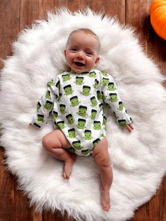 Smiling baby on fluffy white rug, wearing a Frankenstein-themed onesie, surrounded by a cozy autumn setting.