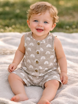 Smiling baby in a rainbow-patterned outfit seated on a blanket outdoors. Bright eyes and cheerful expression.