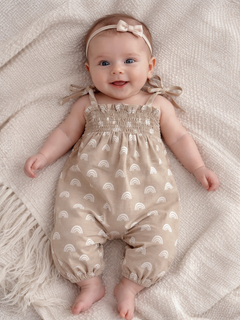 Smiling baby in a beige rainbow-patterned romper, laying on a cozy blanket with a cute headband.
