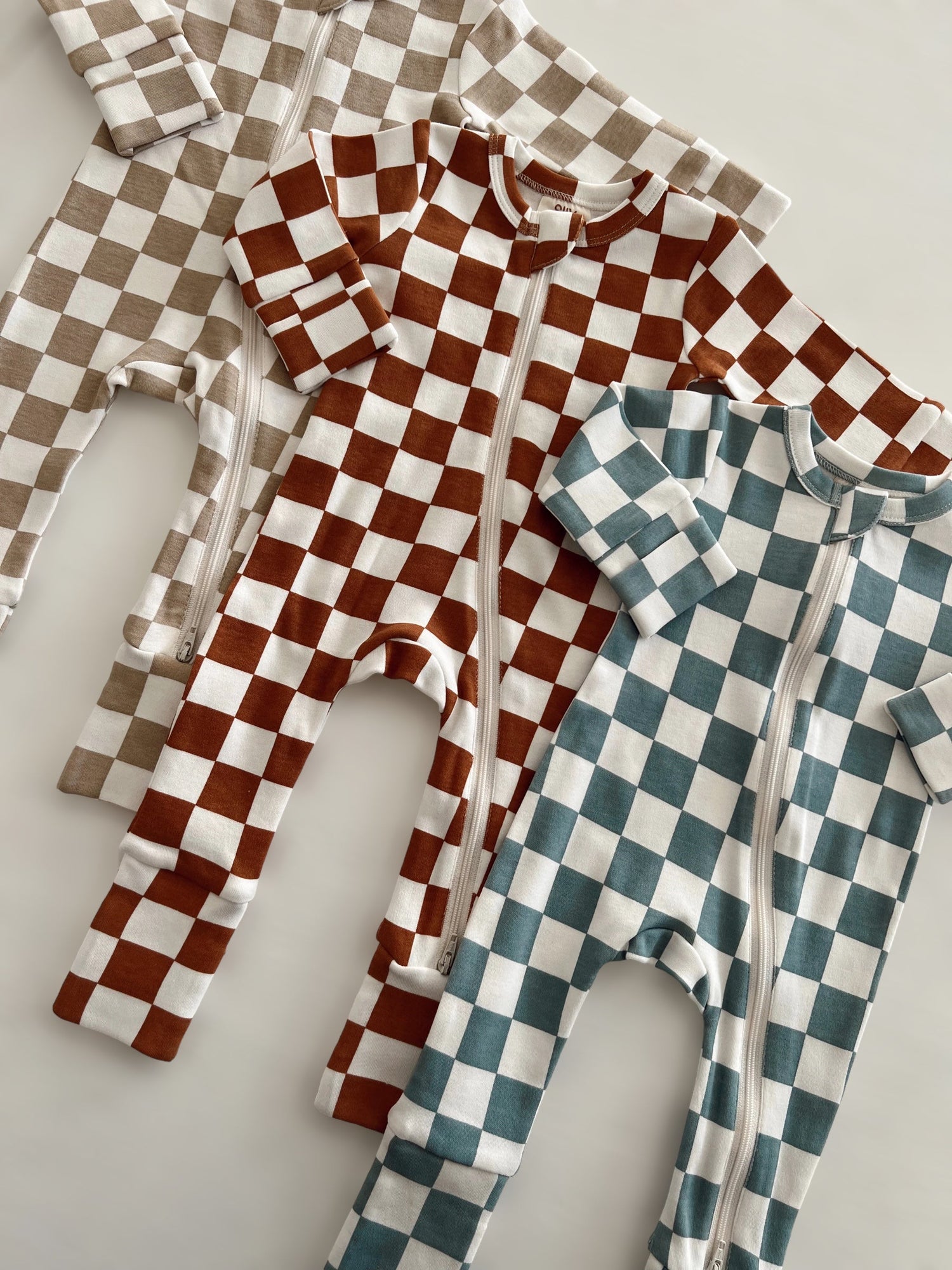 Three checkered baby onesies in brown, blue, and beige laid flat on a light background.