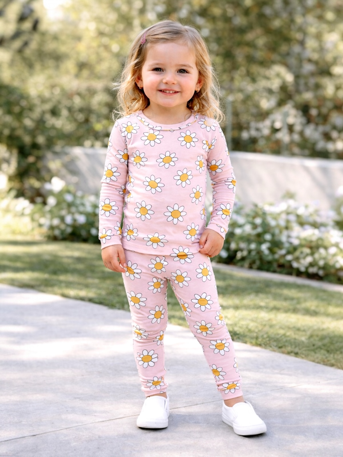 Smiling young girl in pink flower-printed outfit stands outdoors, surrounded by greenery.