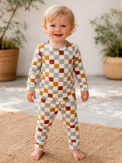 Smiling toddler in colorful checkered pajamas standing on a textured rug with plants in the background.