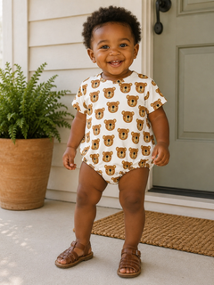Smiling toddler in a bear-patterned onesie stands on a porch, surrounded by greenery and natural light.