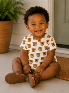Smiling toddler sitting on the floor, wearing a bear-print outfit, surrounded by greenery and a welcoming entrance.
