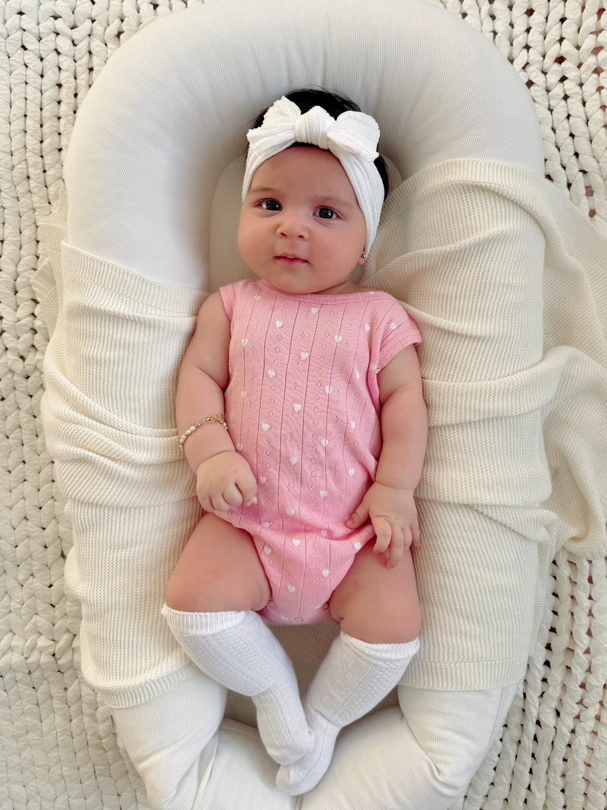 Baby girl in a pink outfit and headband, resting on a soft blanket with a cozy, textured background.