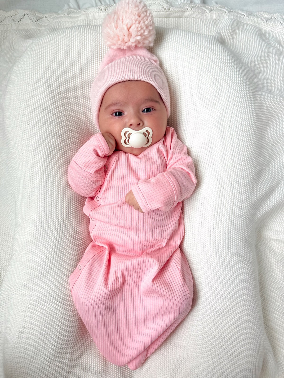 Infant wearing a pink outfit and hat, holding a pacifier, lying on a textured white blanket.