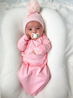 Infant wearing a pink outfit and hat, holding a pacifier, lying on a textured white blanket.
