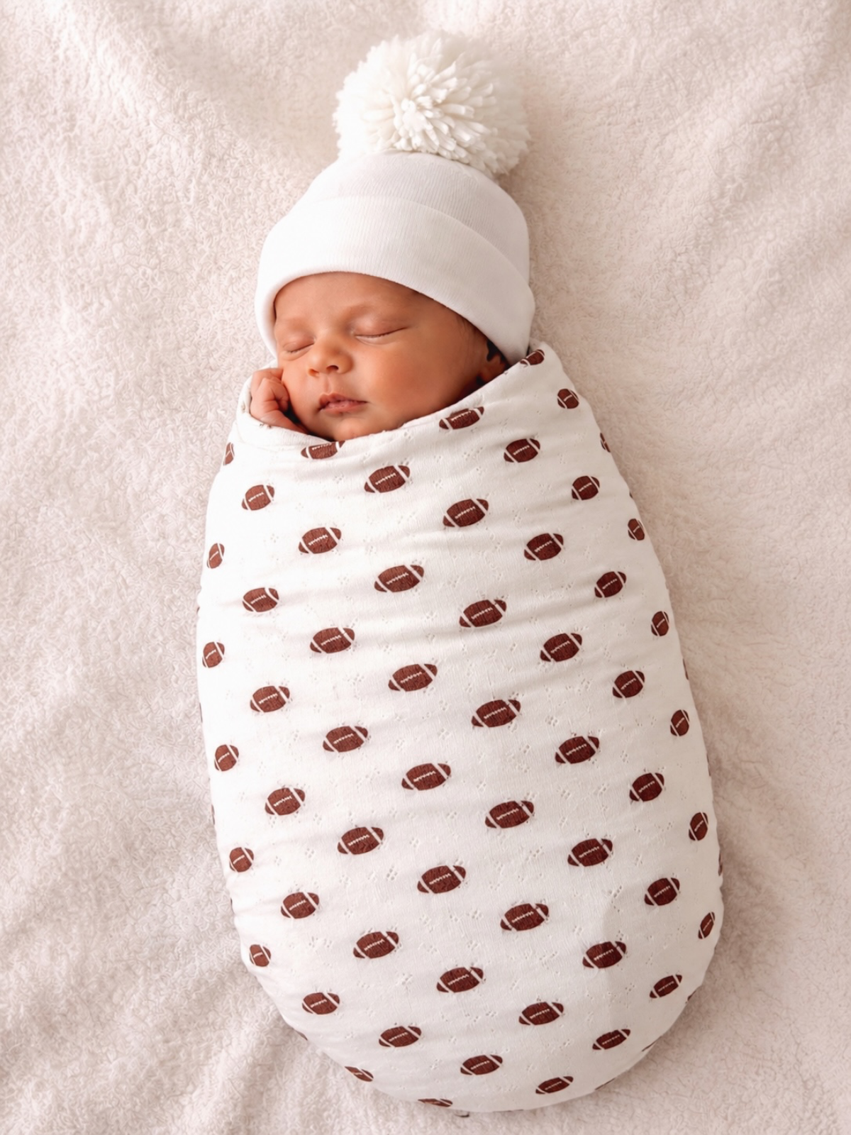 Sleeping newborn wrapped in a football-patterned blanket, wearing a white hat with a pom-pom.