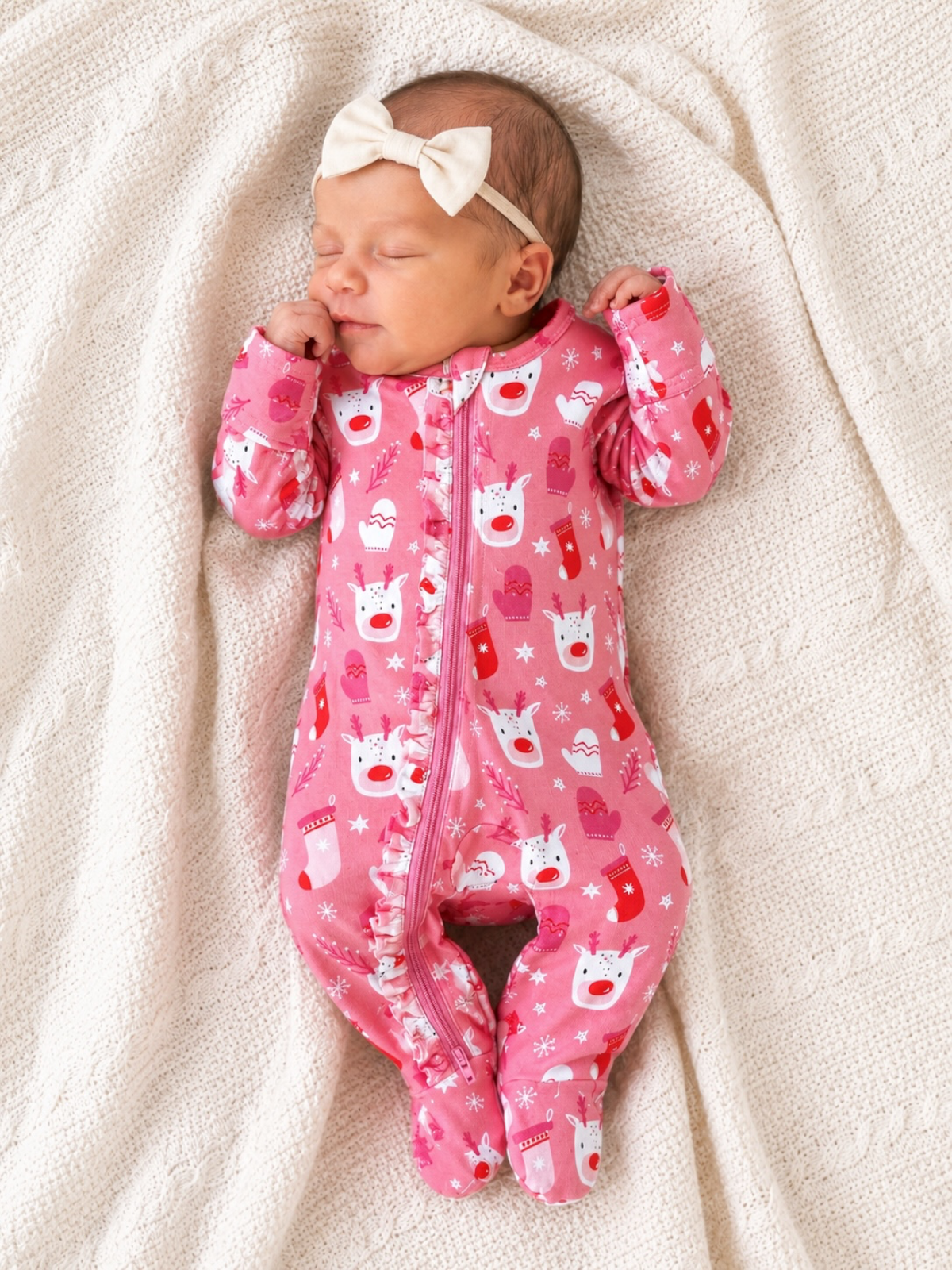 Sleeping baby in a pink reindeer-patterned onesie with a bow headband, resting on a cream blanket.