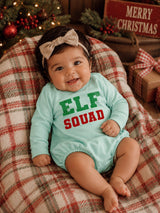 Smiling baby in mint outfit with "ELF SQUAD" text, sitting on plaid blanket near Christmas decor.