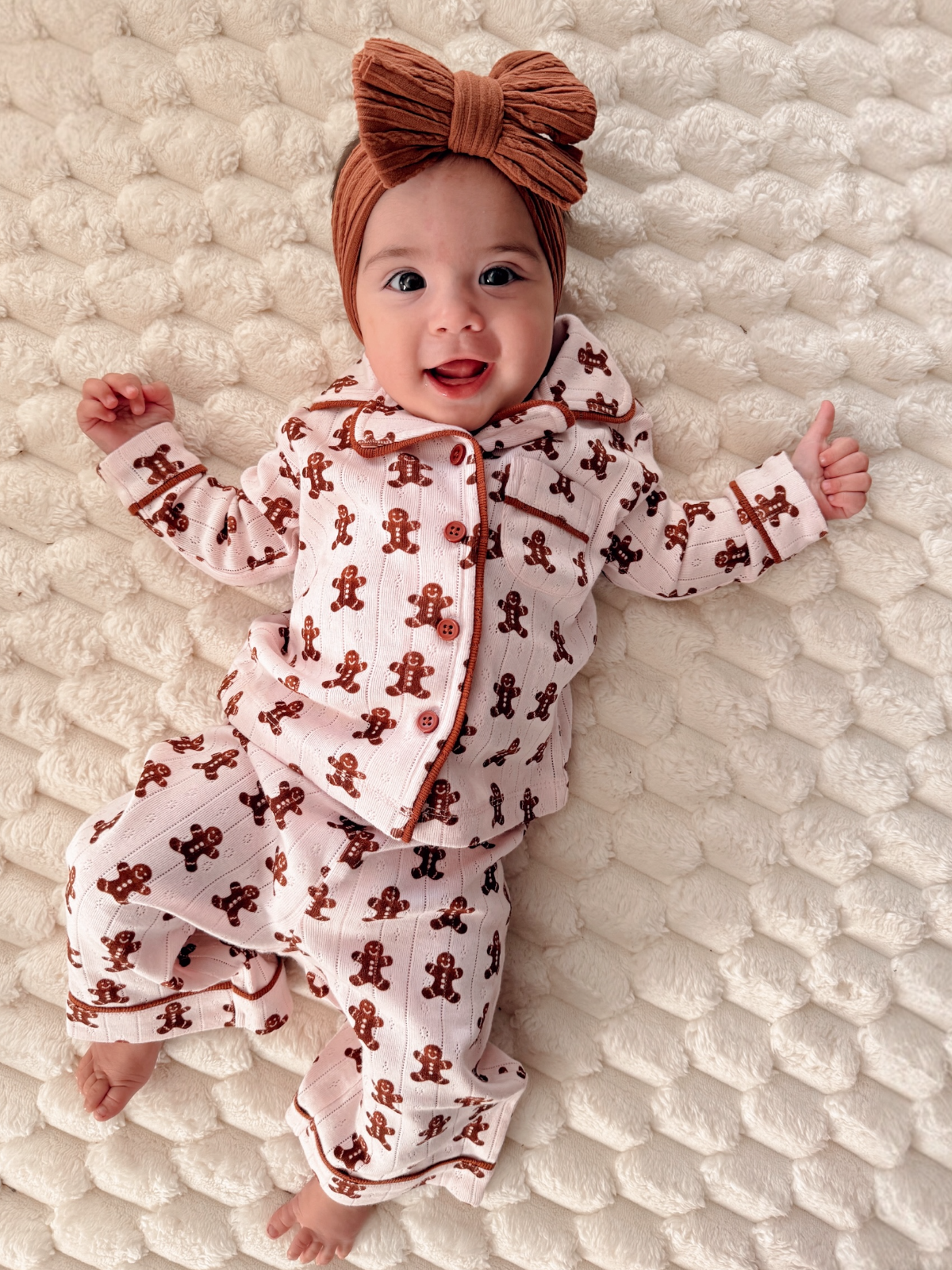 Smiling baby in gingerbread-patterned pajamas on a soft, textured blanket, wearing a large brown headband.