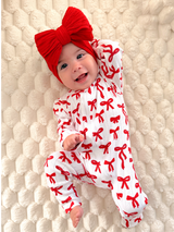 Smiling baby in a red bow headband and patterned onesie on a cozy blanket.