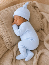 Sleepy newborn in a light blue outfit and hat, nestled on a cozy cream-colored blanket.