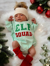 Newborn in an "Elf Squad" outfit with a cream hat and red booties, surrounded by festive greenery and ornaments.