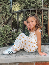 Smiling girl in a sun-printed outfit sitting on a stone wall with greenery in the background.