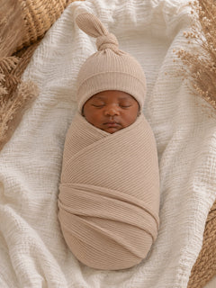 Newborn baby swaddled in a soft beige fabric with a matching hat, resting on a textured white blanket.