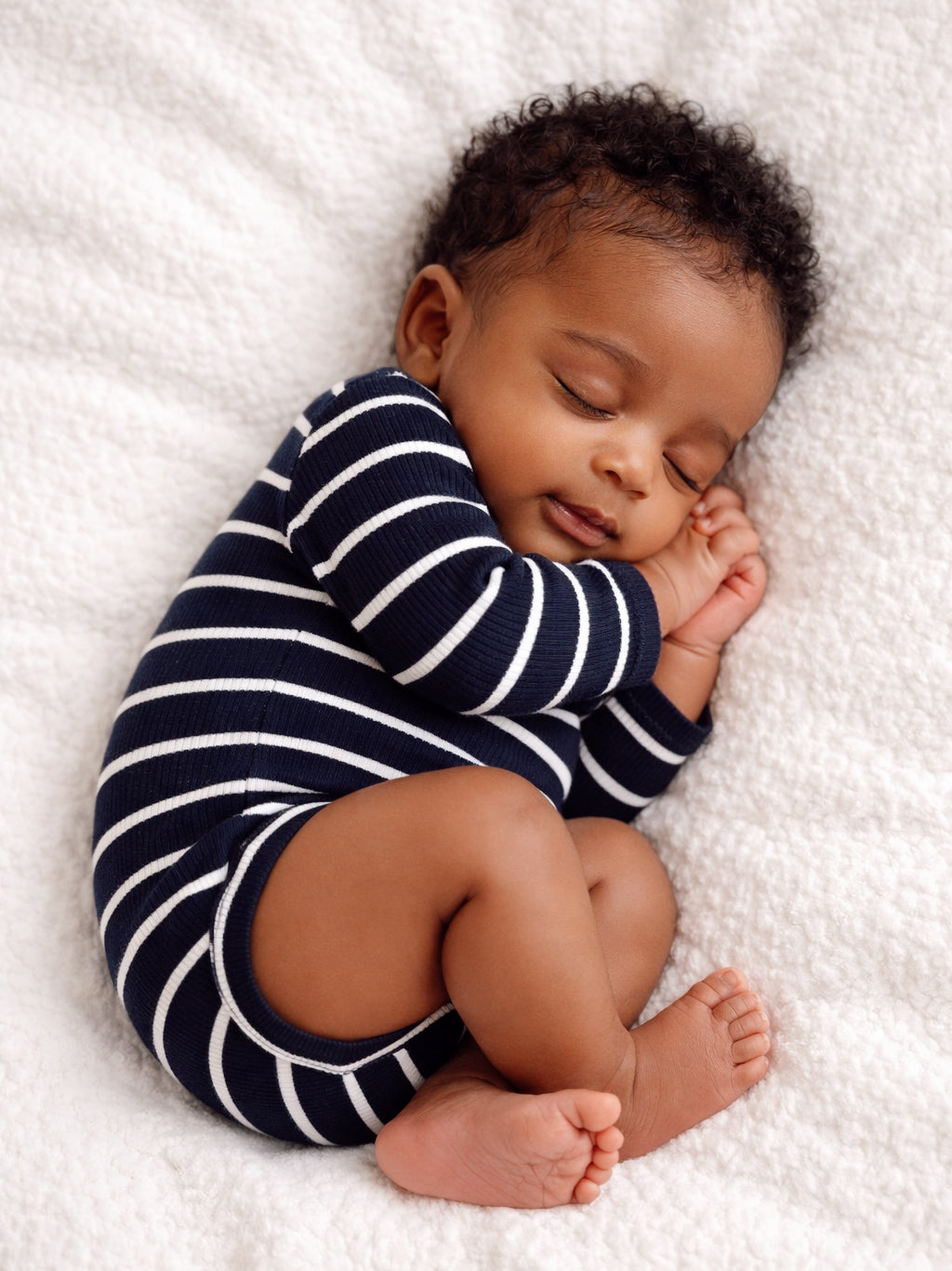 Sleeping baby in striped onesie, curled up on a soft blanket, exuding a peaceful, serene vibe.