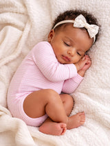 A sleeping baby in a pink onesie rests peacefully on a soft, textured blanket with a lace bow headband.