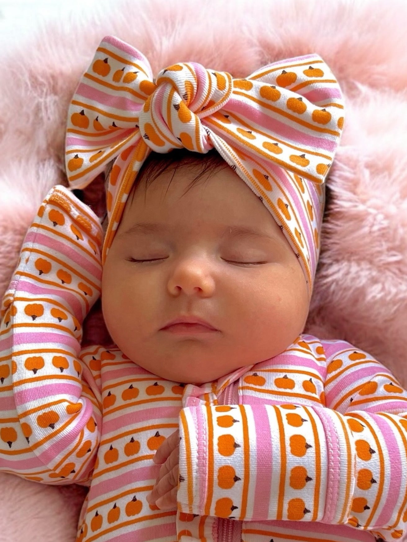 Infant girl asleep with a patterned headband and pajamas on a soft pink blanket.