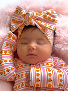 Infant girl asleep with a patterned headband and pajamas on a soft pink blanket.