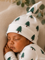 Sleeping baby wearing a white hat and onesie with green tree patterns, peaceful expression, soft background.