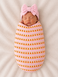Newborn wrapped in pumpkin-patterned fabric, wearing a pink bow, resting on a soft, light-colored blanket.