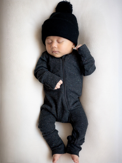 Baby in a black zip-up onesie and pom-pom hat sleeping peacefully on a light-colored surface.