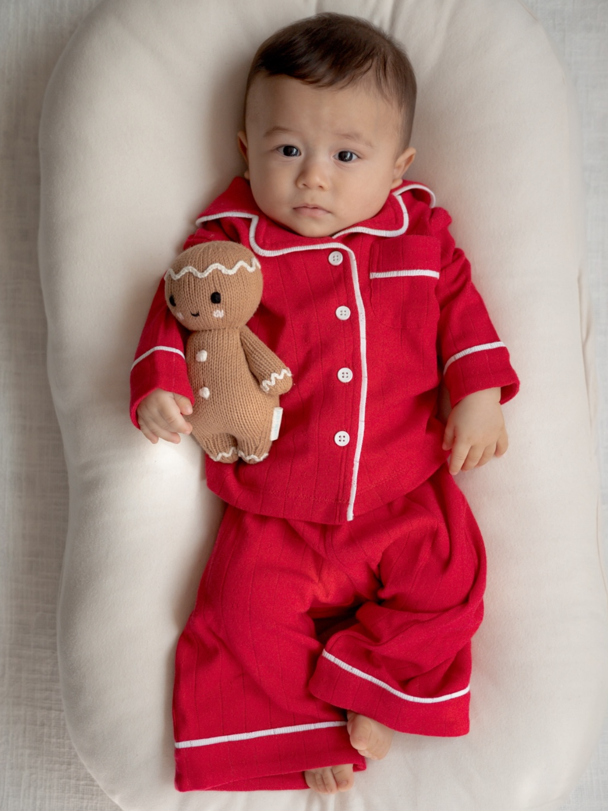 Infant in red pajamas holding a gingerbread doll, lying on a soft pillow.