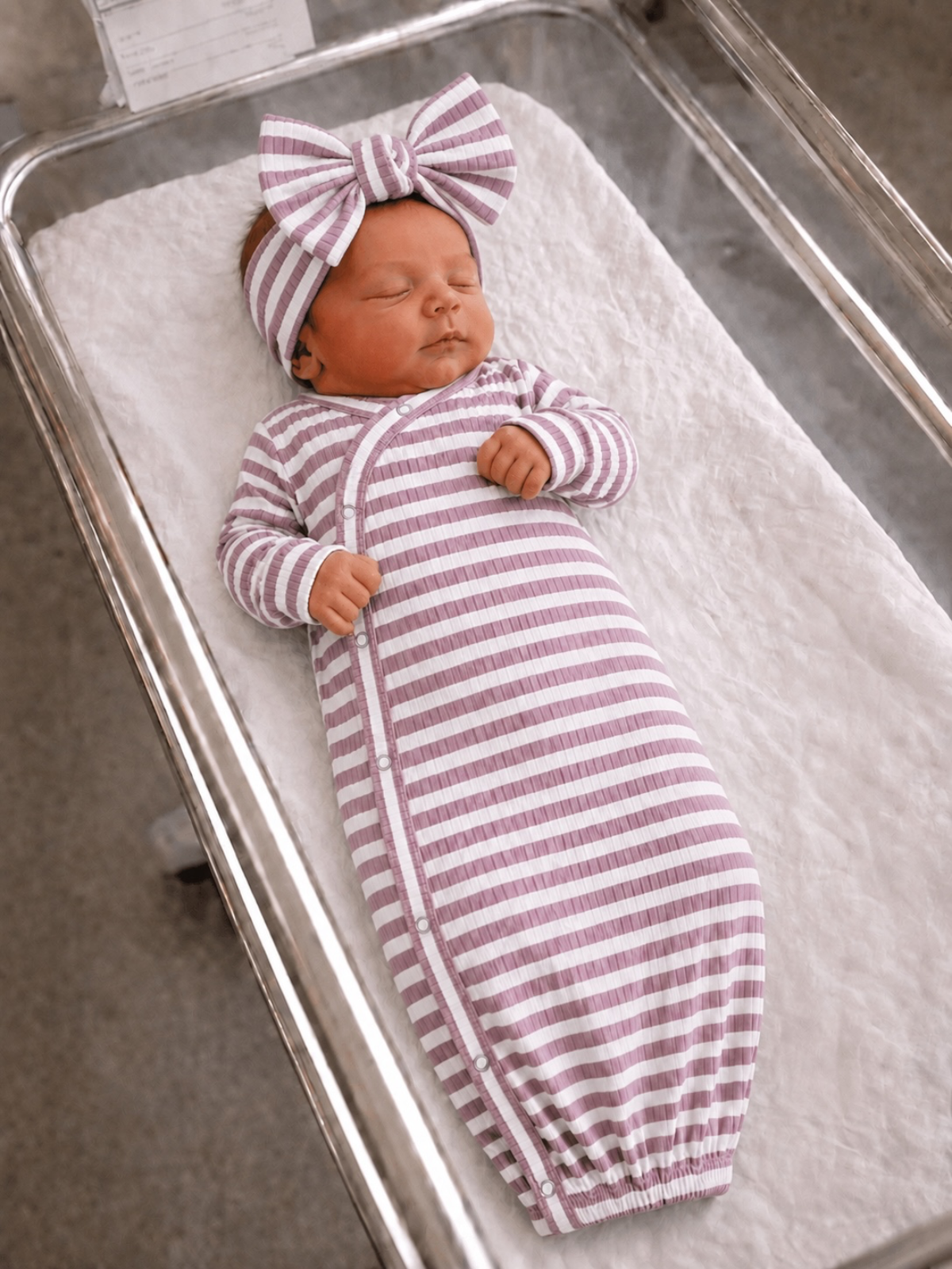 Newborn baby girl in a striped pink swaddle and bow, peacefully sleeping in a clear bassinet.