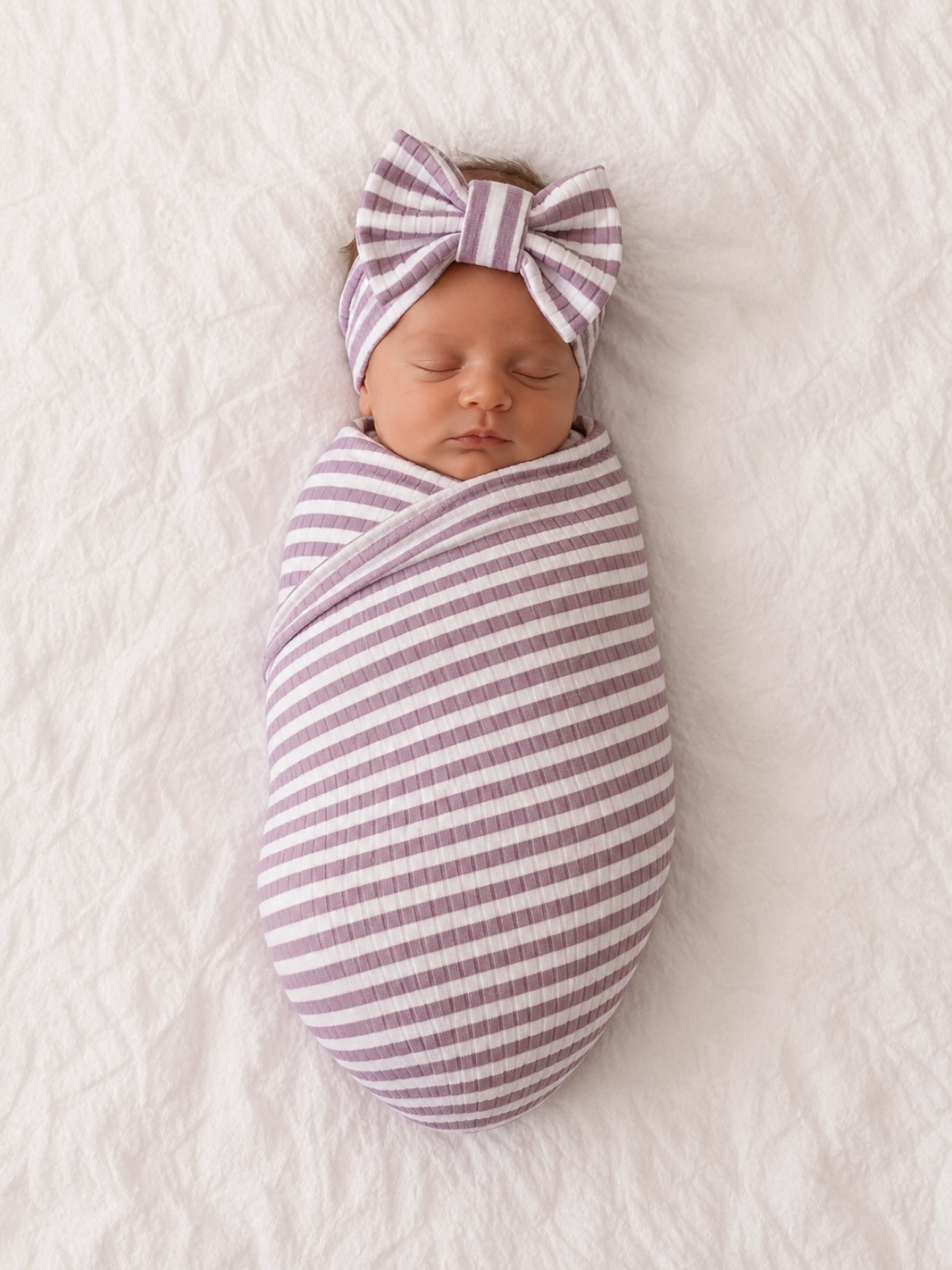 Newborn swaddled in purple striped fabric with a matching bow headband, resting on a soft textured white background.