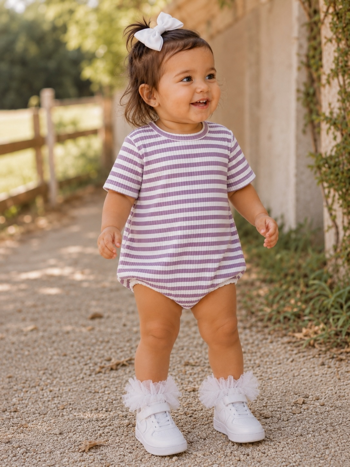 Toddler girl in striped outfit and white sneakers smiles on a sunny path, with greenery and a fence in the background.