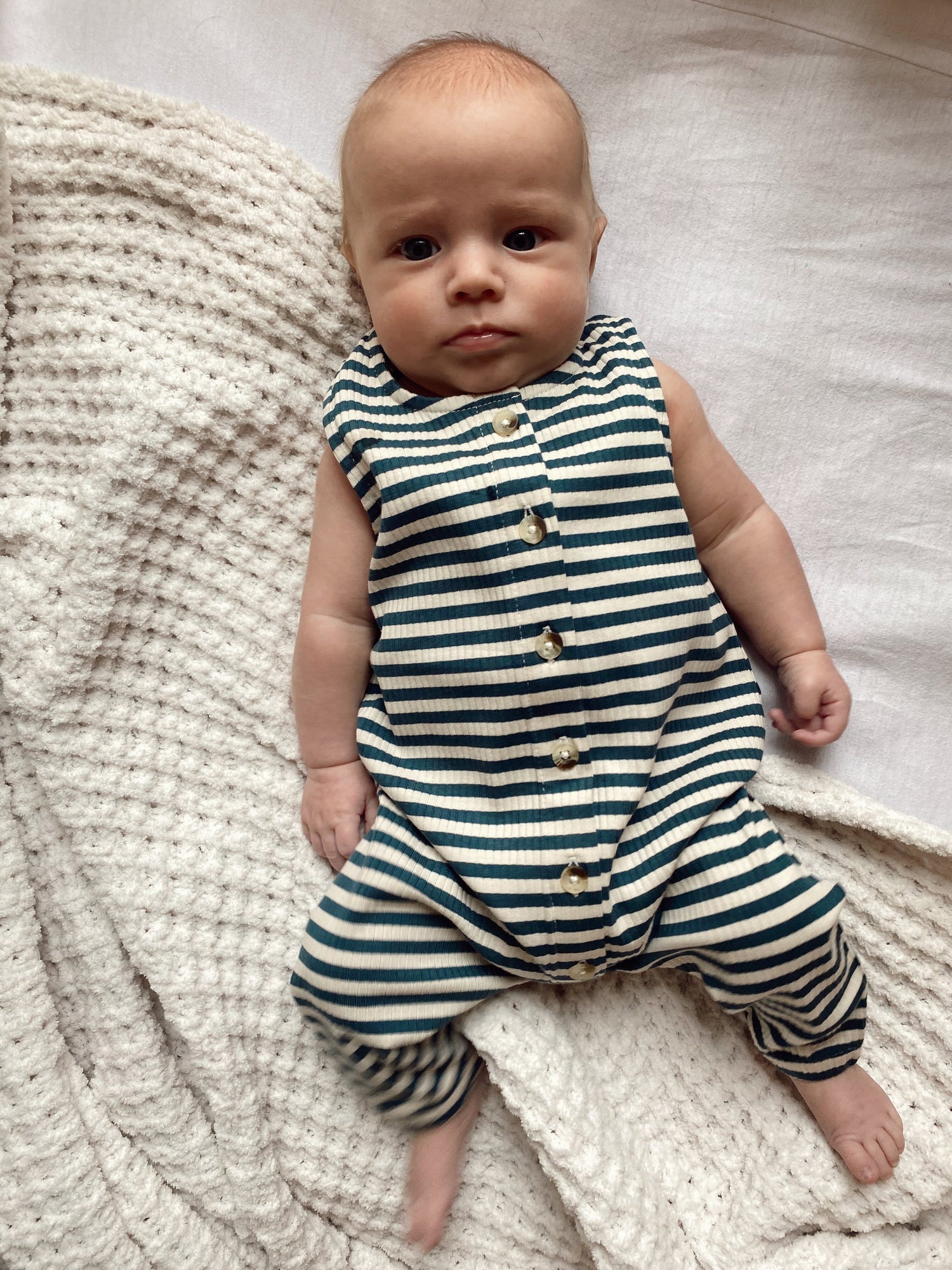 Baby wearing a striped romper sits on a textured blanket, gazing directly at the camera.