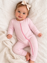 Happy baby in a pink zip-up onesie, with a cream bow headband, lying on a soft white blanket.