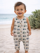 Smiling toddler in a patterned romper stands on the beach, with ocean waves and greenery in the background.