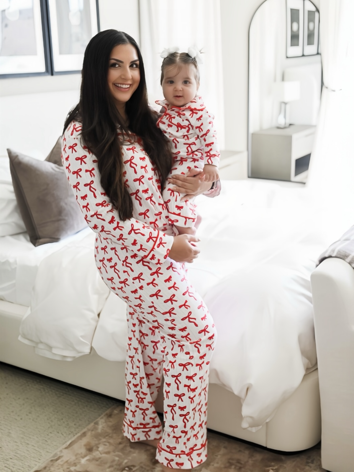 Smiling woman in matching ribbon-patterned pajamas holding a baby, standing near a cozy bed with white bedding.