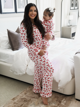 Smiling woman in matching red ribbon pajamas holds a baby in similar outfit, standing in a cozy bedroom.