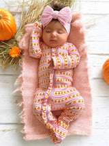 Sleeping baby girl in pumpkin-patterned onesie, pink headband, nestled on soft blanket with pumpkins and flowers.