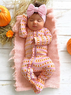 Sleeping baby girl in pumpkin-patterned onesie, pink headband, nestled on soft blanket with pumpkins and flowers.
