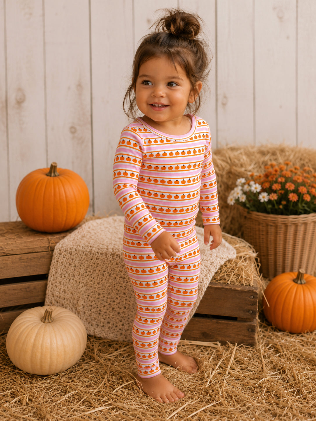 Toddler in pumpkin-themed pajamas smiling among pumpkins and flowers in a cozy, autumn setting.